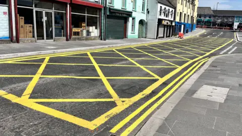 A large yellow box junction leading to traffic lights at a T-junction. Shops line the side of the street.