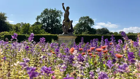 Wander Vision A collection of purple and red flowers in front of an old statue of a man lifting something above his head. There is a large hedge behind it with trees in the distance. The sky above is blue with a few white clouds.