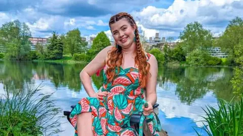 Jenni Pettican Jenni Pettican in her wheelchair by the University of East Anglia lake . She is wearing a red and green watermelon patterned dress and is looking directly at the camera and smiling.