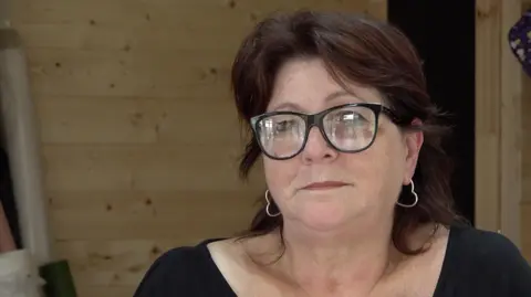 A close-up of Jane Thompson, from the Meridian Action Group. She has dark, shoulder-length hair and wears silver heart-shaped earrings and horn-rimmed glasses with black frames