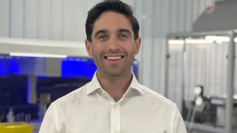 Jeff Bousfield is wearing a white shirt. He is standing in the lab smiling into camera.