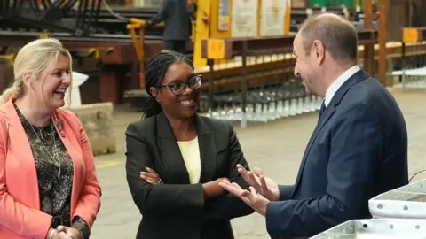 Kemi Badenoch wearing a black suit and yellow t-shirt. She stood in the middle of the frame, smiling and wearing dark-rimmed glasses. Her arms are folded and she is listening to a middle-aged man on the right of the frame wearing a navy suit. The man's hands are folded upwards as if he's demonstrating something to her. On the left of the frame is a blonde woman wearing a pink jacket and dark patterened blouse. She's smiling and listening to what the man is saying. 