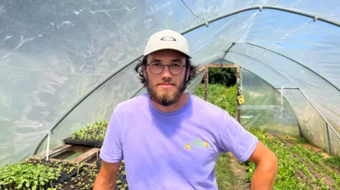 Man wearing purple t shirt stood in a polytunnel filled with growing plants.
