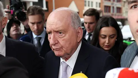 An old man, partially bald with short grey hair, wearing a dark suit and surrounded by a crowd