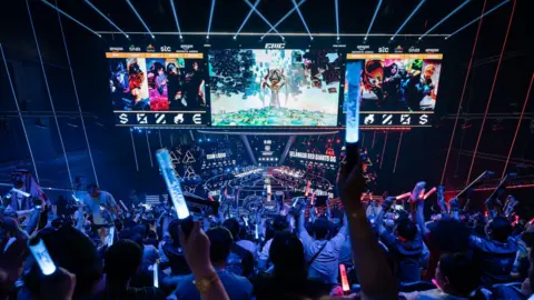The stage at the Esports World Cup has three overhead screens and a large floor with two teams on each side. A seated crowd is seen holding up their arms in support