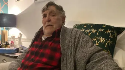 Brian Farmer/BBC Derek Mason: A man wearing a red-and-black checked shirt and grey cardigan sitting in a beige armchair with a black and gold cushion behind him