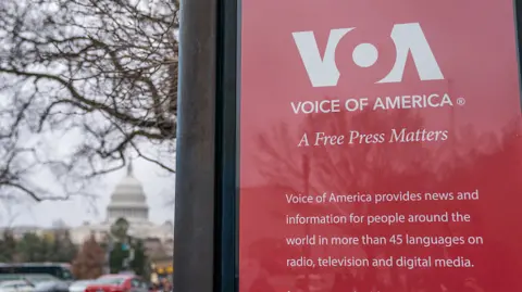 Getty Images menandatangani di luar markas Voice of America di Washington, DC, AS, pada hari Sabtu, 15 Maret 2025. Presiden Donald Trump pada hari Jumat menandatangani perintah eksekutif yang bertujuan mengurangi ruang lingkup delapan lembaga federal sebagai bagian dari kampanyenya untuk merampingkan pemerintah AS, termasuk agen AS untuk media global, yang mengawasi Voice of America. 