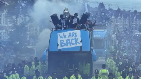 BBC/ Lisa Pitchford An open-topped bus with a banner reading 'We Are Back' is flanked b police officers in hi-viz jackets as it makes its way through a residential street on Coventry. Players on the bus are holding the Championship Trophy alfot at the front of the bus, which is surrounded by smoke from pyrotechnics set off in the crowd.