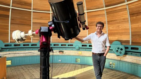 Dr Matt Bothwell standing next to a telescope. He is smiling and wearing a white shirt and black jeans. He is resting his hand on a large telescope. He is standing inside a circular wood-paneled observatory. 