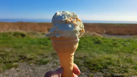 Cumbria lass Barrow in Cumbria. Close up shot of someone holding an ice cream cone with vanilla. They are holding the cone in front of a sea landscape with grass on the bottom half of the photo. The skies are blue. 