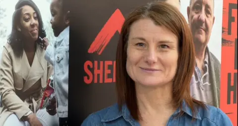 Tracy Guy has shoulder length brown hair. She is wearing a denim dress and is pictured in front of a poster for Shelter. 