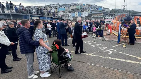 The congregation gathered on the quayside are looking sombre as Helen Lovell-Smith stands at a lectern. Behind her is a RNLI lifeboat.
