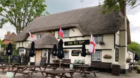 Friends of The Ivy The frontage of the pub takes up the whole photograph - it is painted white with a thatched roof and porch and has a sign saying 'The Ivy Inn'. There are picnic tables outside, with closed parasols. 