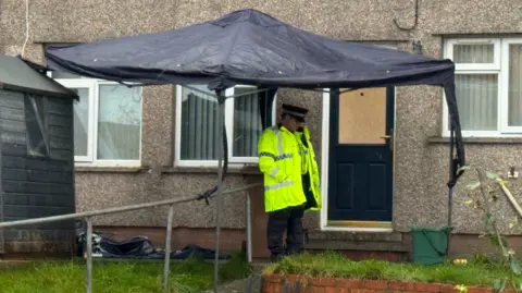 A police officer stands by the door of a home, with a forensic tent visible