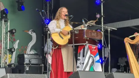 Mari Mathias stands on a stage at the Tafwyl Welsh-language festival. She sings as she strums a guitar. She wears a long red skirt, a beige waistcoat and a white billowy with a cream apron.
