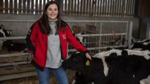 Coleg Sir Gâr Sara Roberts, 22 - agriculture lecturer at Coleg Sir Gâr - standing with a calf in a cattle shed, wearing a red coat over a grey jumper and jeans.  