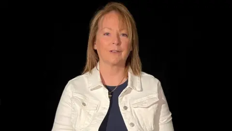 BBC Monica Johnson has light brown hair of shoulder length and is wearing a white jacket above a dark blue. It has a gold necklace with a cross.