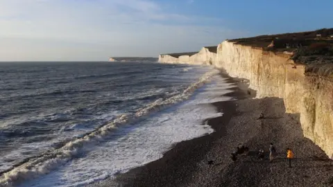 People are walking on the beach below the cliffs at Birling Gap. The shingle is heaped up on the shoreline and the waves are coming in at an angle. The beach runs under a stretch of cliffs with fields on the cliff tops and a row of houses.
