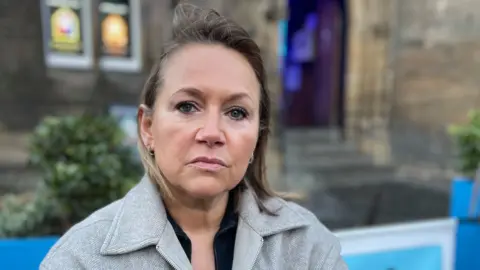 Louise has blonde hair. She is wearing a black top and a grey coat. There is a pub in the background.