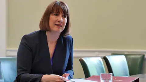 Reuters Rachel Reeves sits at a desk with empty chairs beside her. She's leaning forward and appears to be listening and is holding a piece of A4 paper