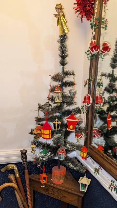 Lisa Watts A skinny green tree with a gold angel on top, hung with a variety of retro decorations including red and gold lanterns.