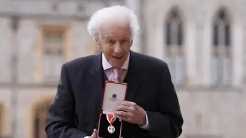 PA Media An elderly man with white hair wearing a suit, showing off his medal 