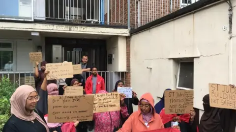 A group of woman stand outside a building holding cardboard signs that read slogans such as 'St Jude's Buildings Not Safe'.