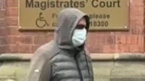 Man wearing a grey coat with a hood over his head walking outside a court wearing a face covering and dark glasses.