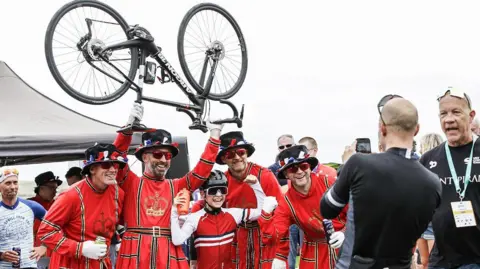 Stuart Boulton Four men in Beefeater costume holding aloft a bicycle, and a young cyclist with raised arms in front of them. A man in lycra cycling wear takes a photo of them on a mobile phone while other people look on