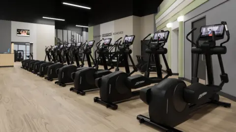 Everyone Active A large interior room with a wooden floor and walls in grey, beige and green. A row of black cross trainers runs from one end of the room to the other