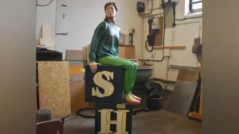 A restored sculpture shows a seated human figure positioned on two stacked black blocks with large cream‑coloured letters “S” and “H.” The sculpture is placed inside a workshop with tools, cables, wooden panels and machinery visible in the background. The figure wears a long‑sleeved top, trousers and shoes.