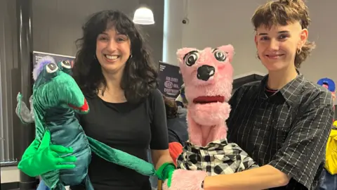 A woman with dark colour hair and a black top holds a green, google-eyed puppet, she stands next to a person with short curly hair wearing a check shirt and holding a pink, bear-like puppet. 