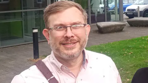 Mike Isherwood A man with blonde hair, glasses and a beard, wearing thin-framed glasses and a pale pink shirt standing in an area partly paved and partly covered in grass, in front of a glass-fronted building.