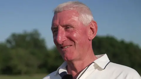 Nic Downes has short white hair and is smiling whilst looking off-camera. He is wearing cricket whites.