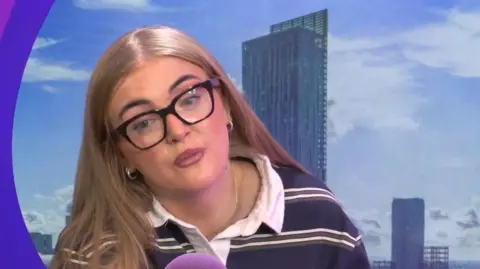 Lauren Jean, who has long blonde hair and is wearing black framed glasses, speaks into a purple microphone.