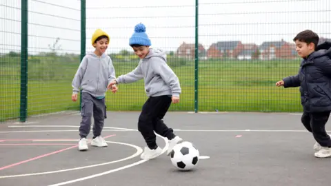 Three young boys play football inside a fenced games area, houses in the background
