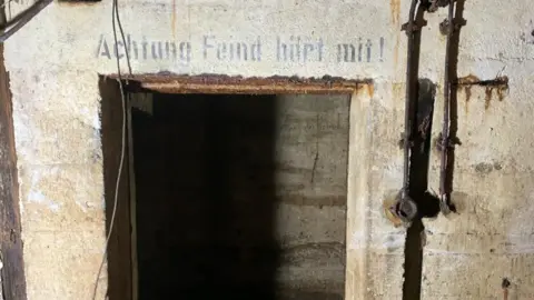Shaun Tullier The German phrase "achtung feind hort mit" - which translates to "beware, the enemy is listening" - written in black letters on the wall of a bunker found underneath a house in Guernsey.