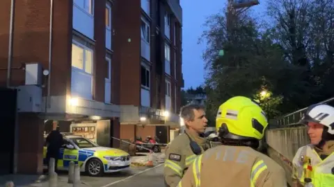 Firefighters stand to the right of the image talking, while police tape surrounds the block of flats and a police car parked under the block