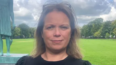 BBC Jackie has glasses on top of her head. She has shoulder length brown hair. There's a statue on one side of her and a field behind her.