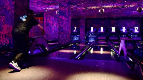 Lane 7 A man in a bowling alley, rolling a ball. The room is dark, with purple colours, with glitter balls ahead, and a number of numbered lanes. The man is dressed in black with white trainers on. 