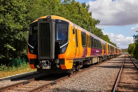 An orange and black Class 730 electric train can be seen on a track with trees either side of it. 