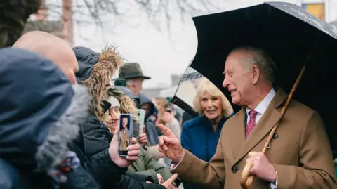 King Charles is in a smart brown double-breasted coat, with a red tie. He is holding a black umbrella over his shoulder. Queen Camilla is behind him in a blue coat. Charles is smiling or laughing while facing someone in the crowd. It is overcast above.