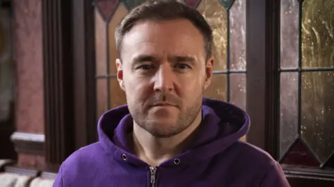 ITV/Coronation Street Tyrone Dobbs played by Alan Halsall wears a purple hoodie and is staring into the camera lens with a straight face.