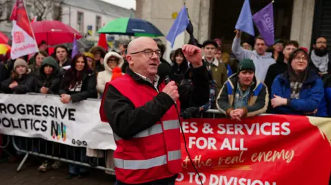 PA Media A man in a red hi vis vest speaking into a microphone outside in front of a group at a counter protest. The group are behind a barrier and they are wearing coats, and holding umbrellas and flags.