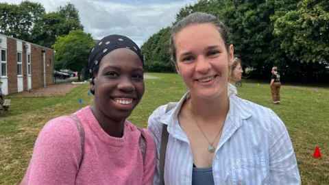 The image shows two girls standing close together with a field behind them. One is in a pink jumper and the other is in a blue and white stripy shirt. There is a building on the back left of the picture. 
