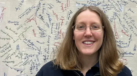 Gemma Clarke - a woman with brown hair - smiles as she stands in front of a document covered with handwritten signatures in various colours. She is wearing oval glasses and a blue cardigan.