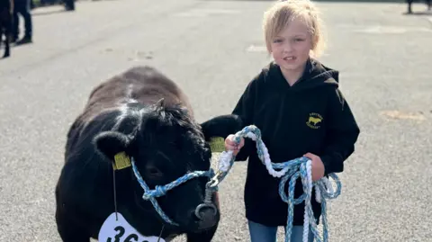 Sarah-Jane is short and has blonde hair which is tied back. She is holding onto a small black cow with a blue lead. Sarah-Jane is wearing a black hoodie, blue skinny jeans and brown boots. The cow has yellow tags in its' ears and has a white number hanging from its neck.
