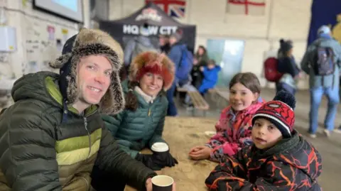 Andrew, Victoria, Saffron and Finn all sat at a table in the hanger. They are all smiling at the camera.