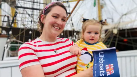 Woman in white and red striped top and daughter holding an RNLI flag, with tall ships in the background.