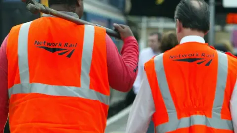 A stock image of two Network Rail workers are walking along a train platform with their backs to the camera. They are both wearing hi-viz orange vests with company branding. A train can be seen behind them.
